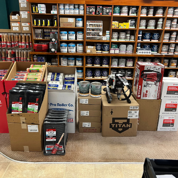 Shelves filled with cans of paint and painting supplies, including brushes and rollers, organized neatly in a store. Boxes and tools are displayed below.