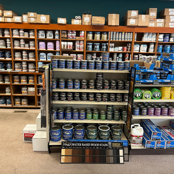 Store interior with shelves displaying an array of paint cans and supplies. A prominent display features water-based wood stain varieties. The setting appears organized and vibrant.