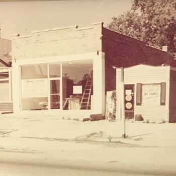 Vintage photo of a small brick storefront with large display windows and adjacent wooden building, evoking a nostalgic, small-town atmosphere.