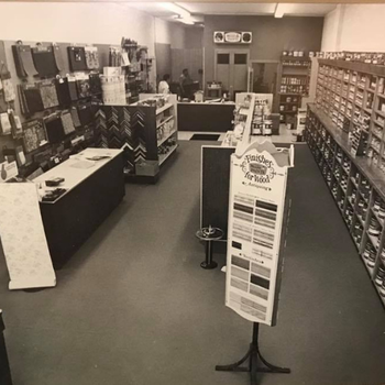 A vintage black-and-white photo of a home improvement store interior, featuring shelves of supplies, a "Finishes for Wood" display, and a counter.