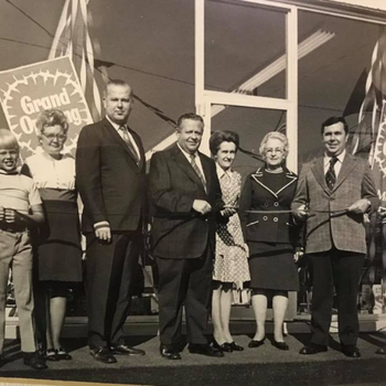 A vintage black-and-white photo shows a group of seven people, including a child, lined up in front of a building with "Grand Opening" signs, smiling.