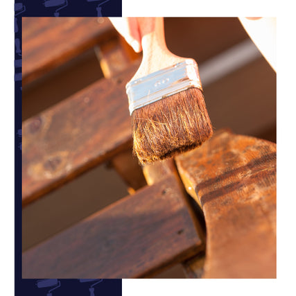 Interior StainsA hand holding a paintbrush is applying varnish to a wooden surface in bright sunlight, highlighting the rich wood grain and warm tones.